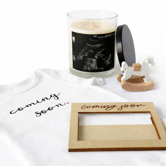 Candle with ultrasound image, 'coming soon' t-shirt, and wooden frame on a white surface
