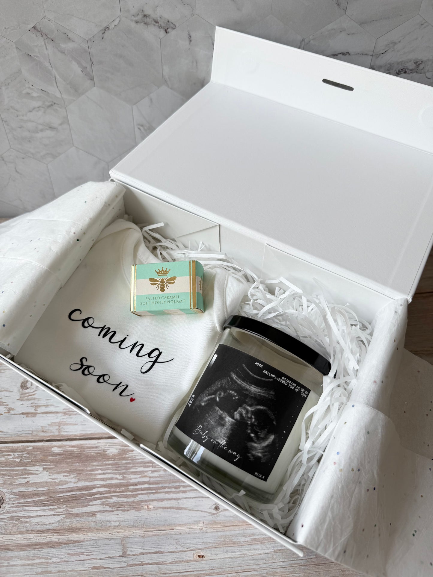 Gift box with 'coming soon' shirt, small box, and ultrasound photo on a marble surface