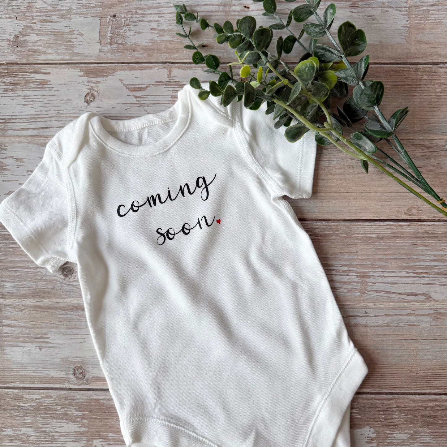 White baby onesie with 'coming soon' text on a wooden surface with greenery.
