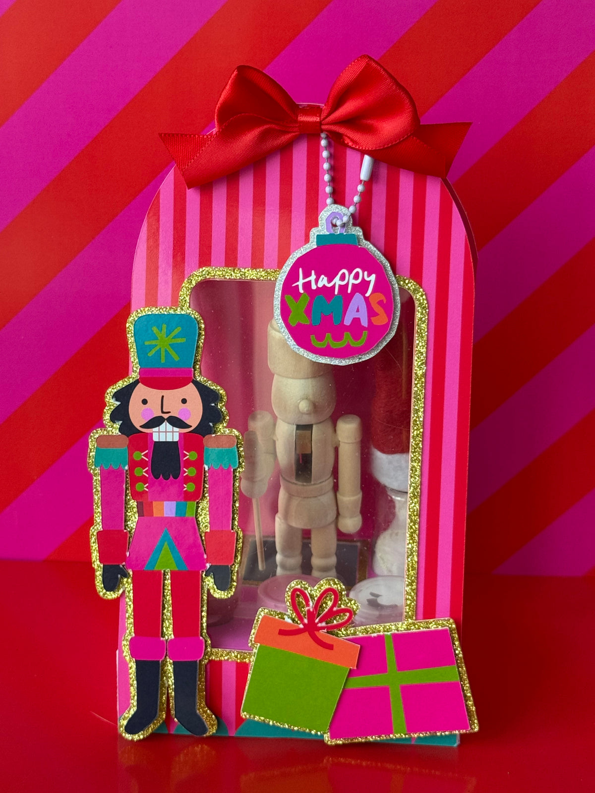 A festive Christmas gift box with a red ribbon bow and a clear window displaying a wooden nutcracker. A colorful 'Happy LMAO Christmas' ornament tag hangs from the top, and a die-cut nutcracker figure and gift boxes are placed in front, all set against a bold pink and red striped background.