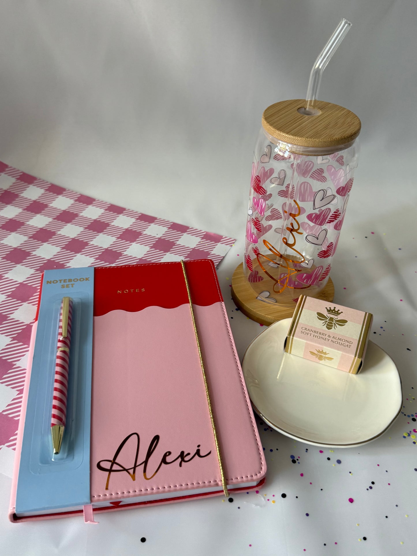 Pink notebook with 'Alex' on the cover, pen, and floral-patterned tumbler on a checkered tablecloth.