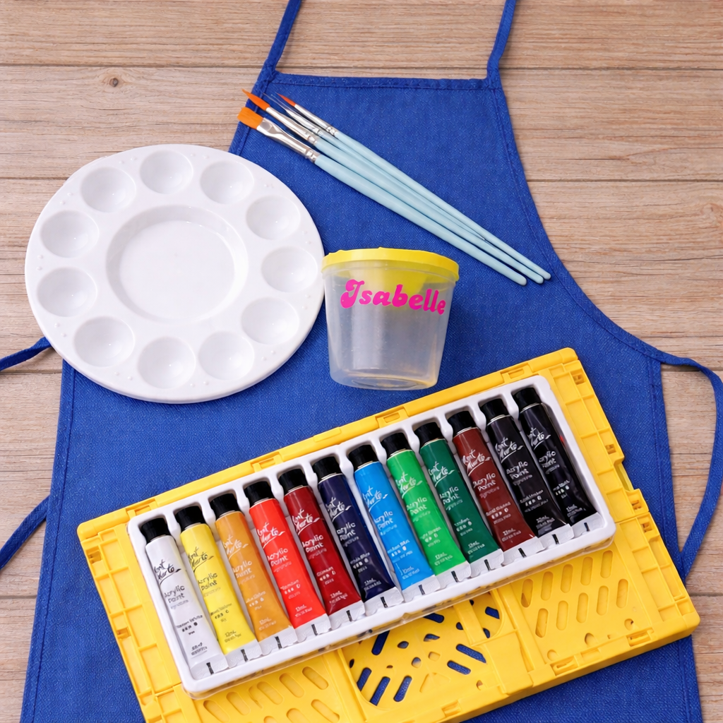 Set of watercolor paints in a yellow tray with an apron, paintbrushes, and a palette on a wooden surface.