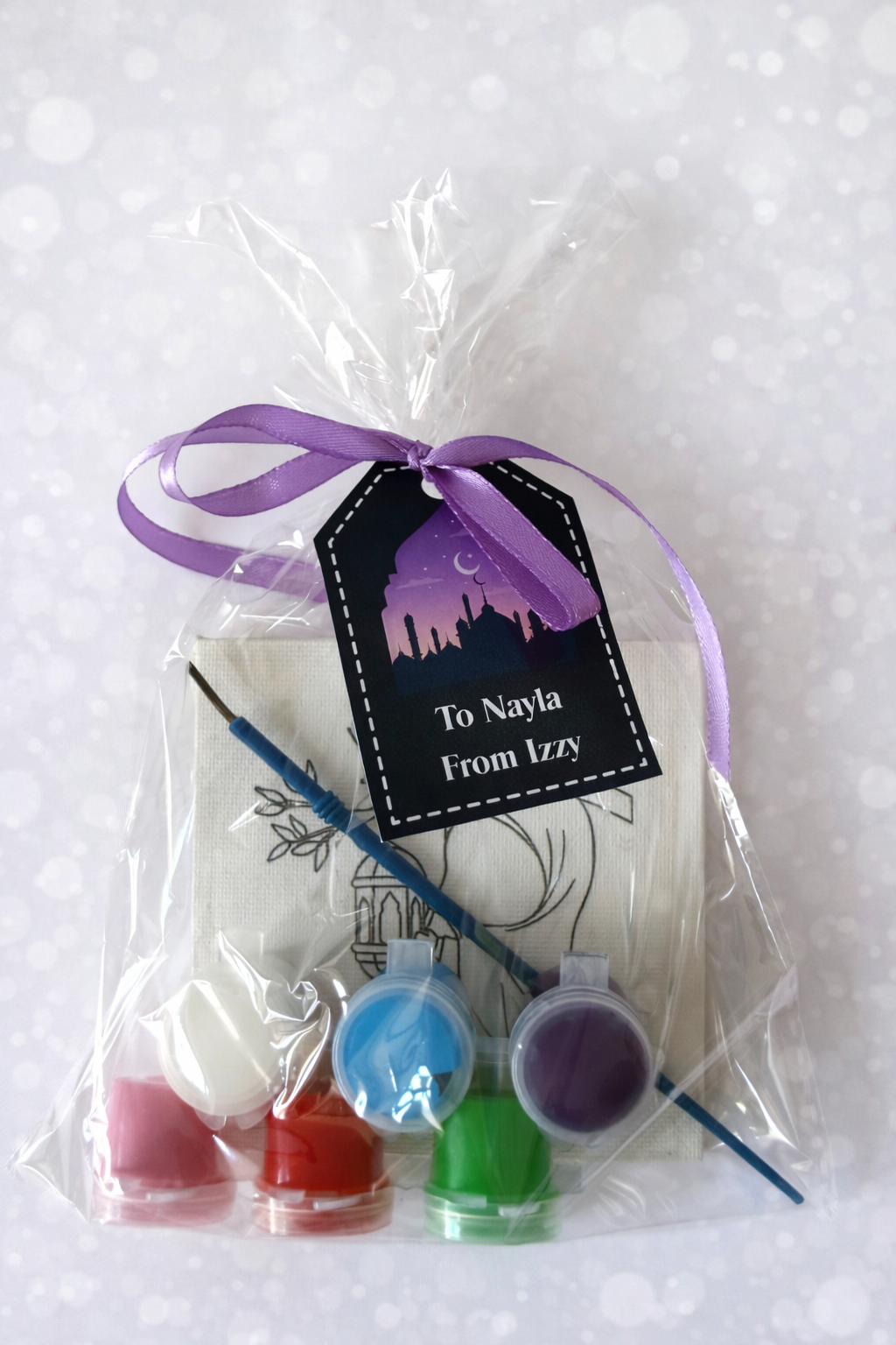 Packaged art supply set with paint bottles and brushes, tied with a purple ribbon and personalized tag.