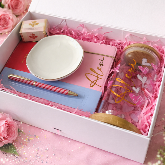 Gift set with a bowl, pen, and mug in a box on a pink surface with flowers.
