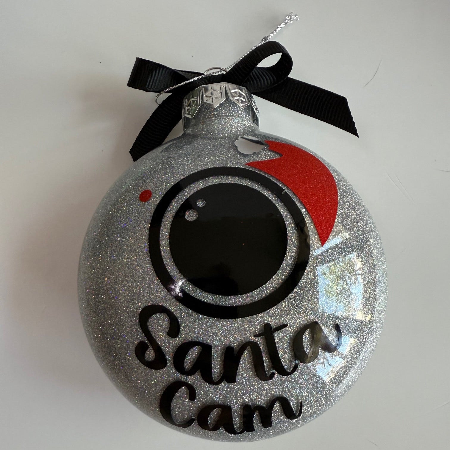 Decorative ornament with a black bow, red dot, and 'Santa Cam' text on a white background