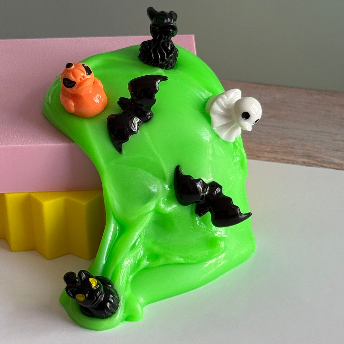 A bright neon green slime-like product with small Halloween-themed figurines, including black bats, a white ghost, and an orange pumpkin, displayed on a pink and yellow block against a neutral background.