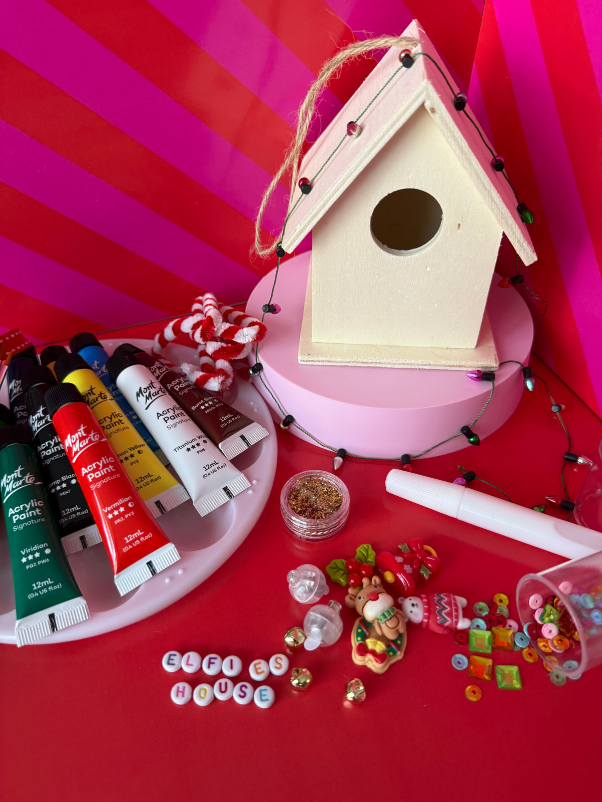 A wooden birdhouse ornament displayed on a pink pedestal with craft supplies around it, including paint tubes, beads, sequins, and small decorative items, against a bright pink striped background.