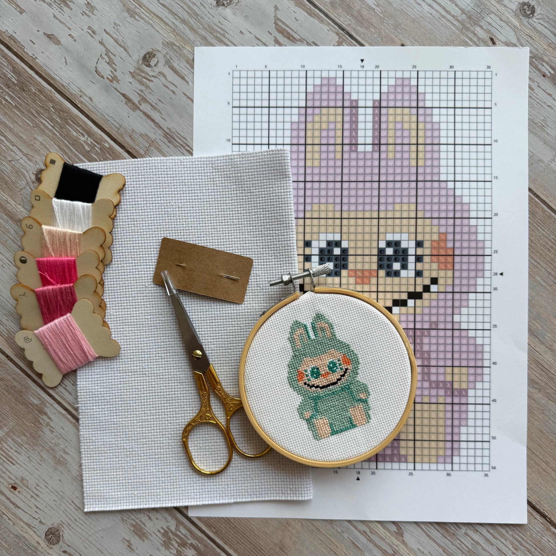 Cross-stitching materials including a pattern sheet, hoop with partially completed design, scissors, and fabric on a wooden surface.