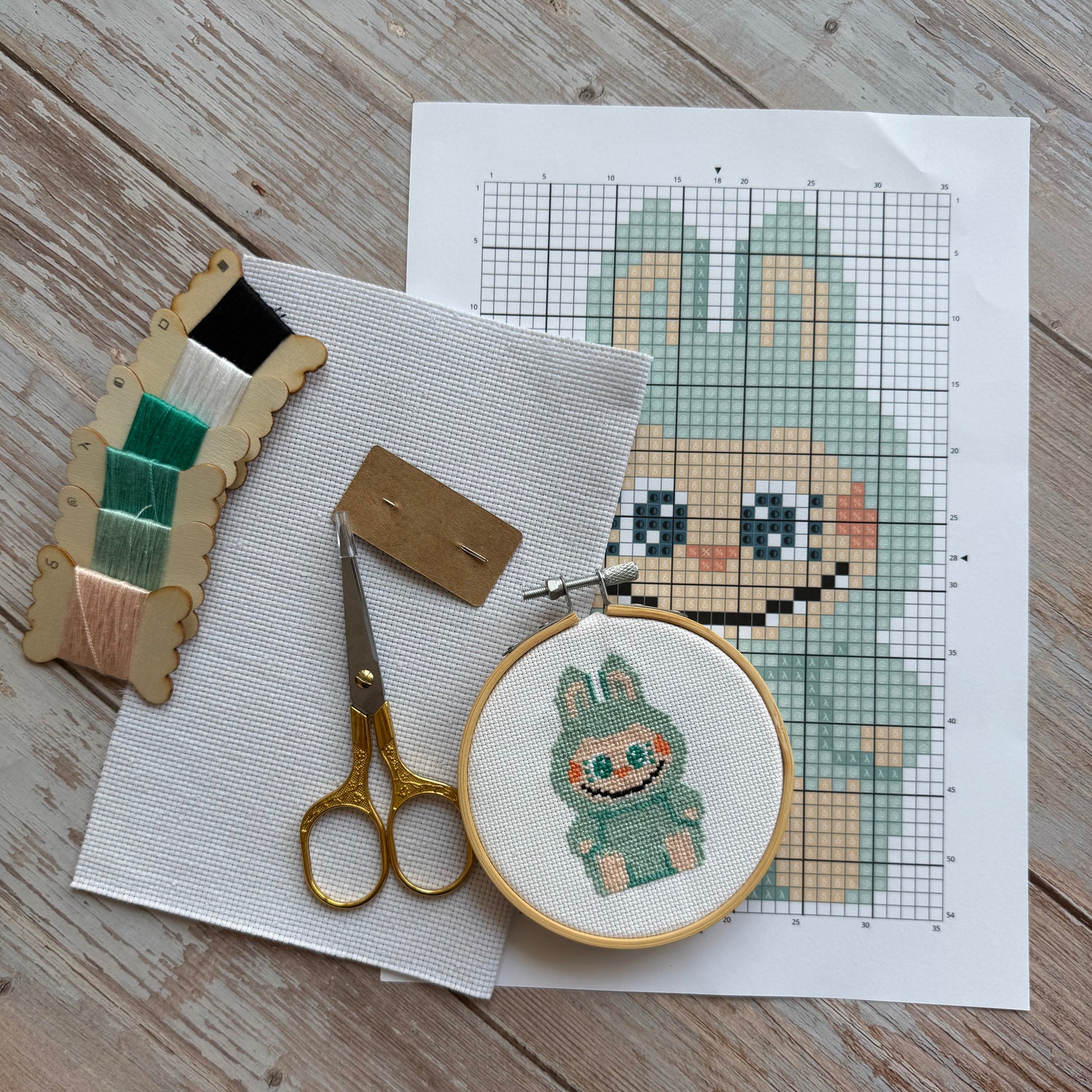 Cross-stitching materials including fabric, hoop, scissors, and pattern on a wooden surface.