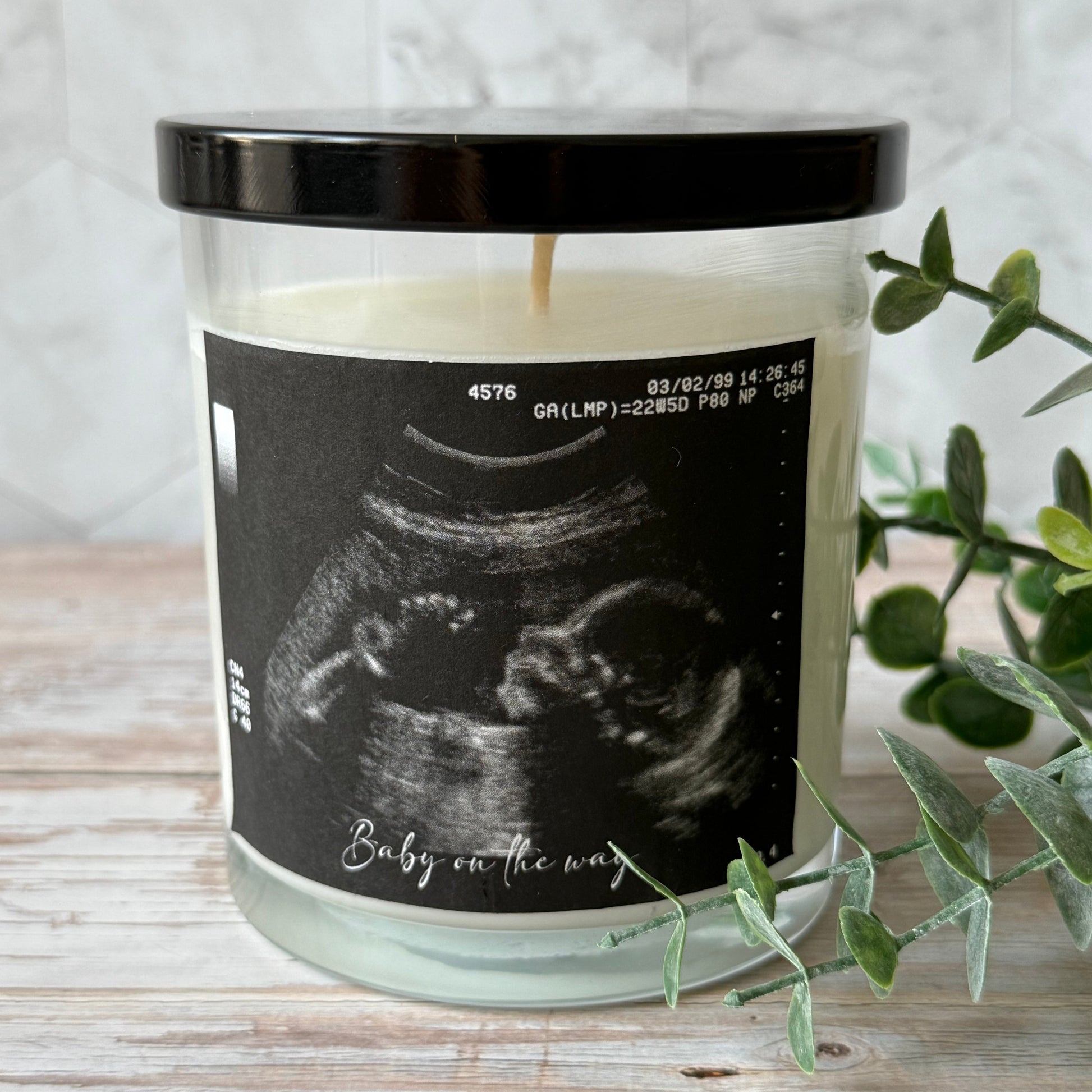 Candle with ultrasound image and 'Baby on the way' text on a wooden surface with a plant.