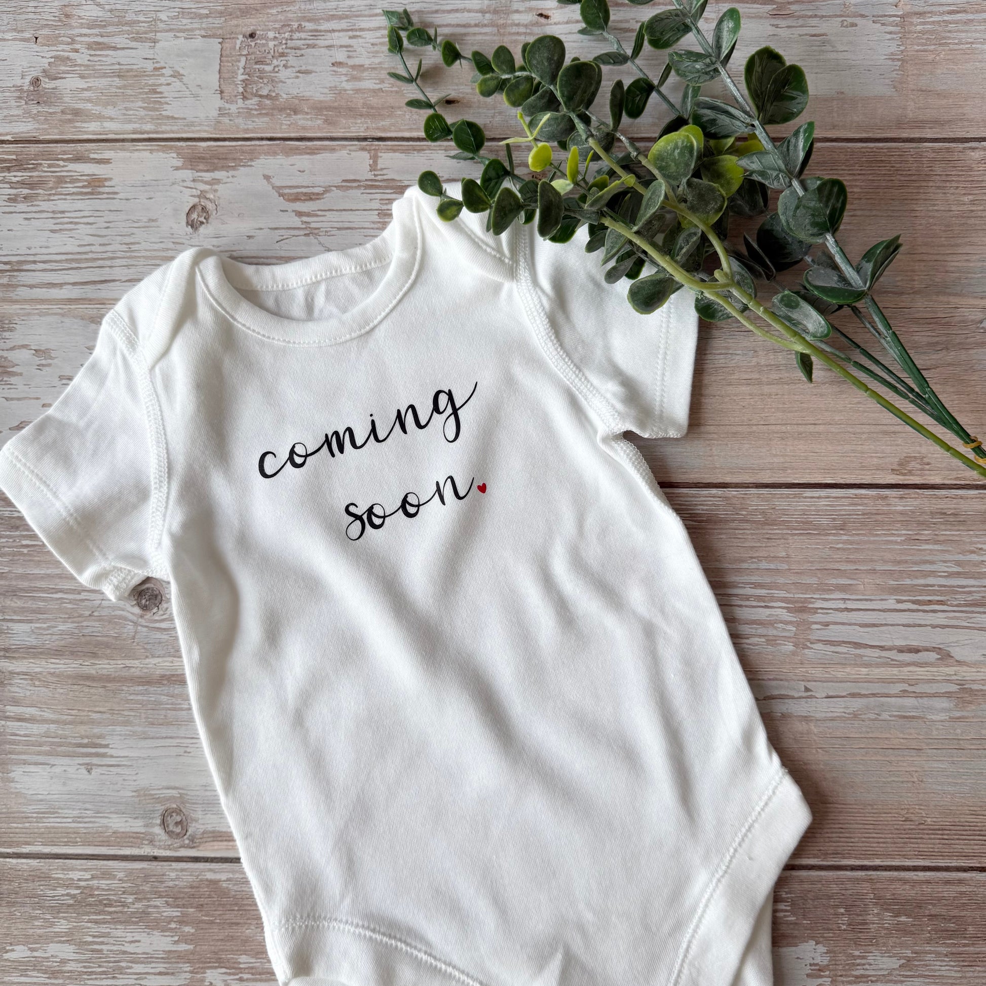 White baby onesie with 'coming soon' text on a wooden surface with greenery.
