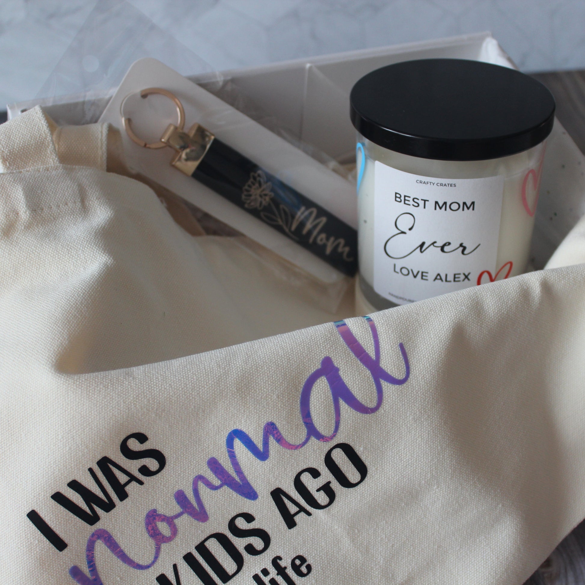 A gift box set containing a personalized candle, a canvas tote bag with a printed quote, and a keychain, all displayed on a wooden surface.