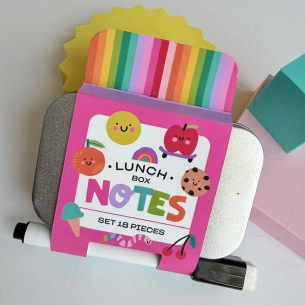 A pink Lunch Box Notes set with 18 pieces, featuring cute cartoon illustrations of fruits, a rainbow, and a smiling sun. The notes are displayed in a lunchbox-shaped holder on top of a gray metal tin, with a white marker resting in front, set against a clean white background.