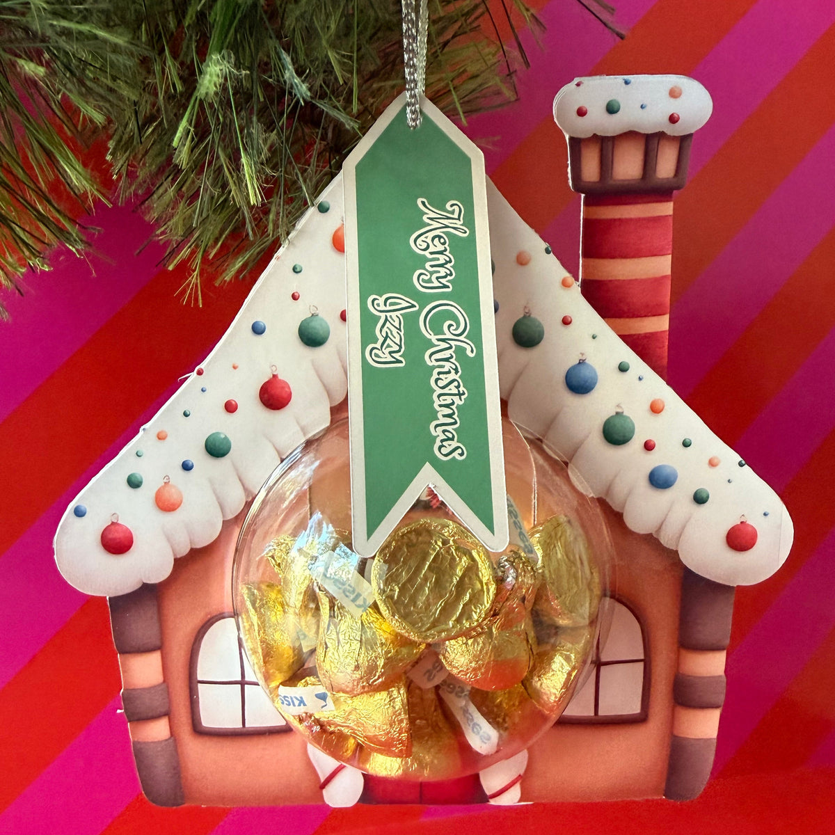 A gingerbread house Christmas ornament filled with foil-wrapped chocolates, featuring a green hanging banner that reads 'Merry Christmas' and colorful candy decorations, shown against a festive red and pink striped background with pine branches at the top.