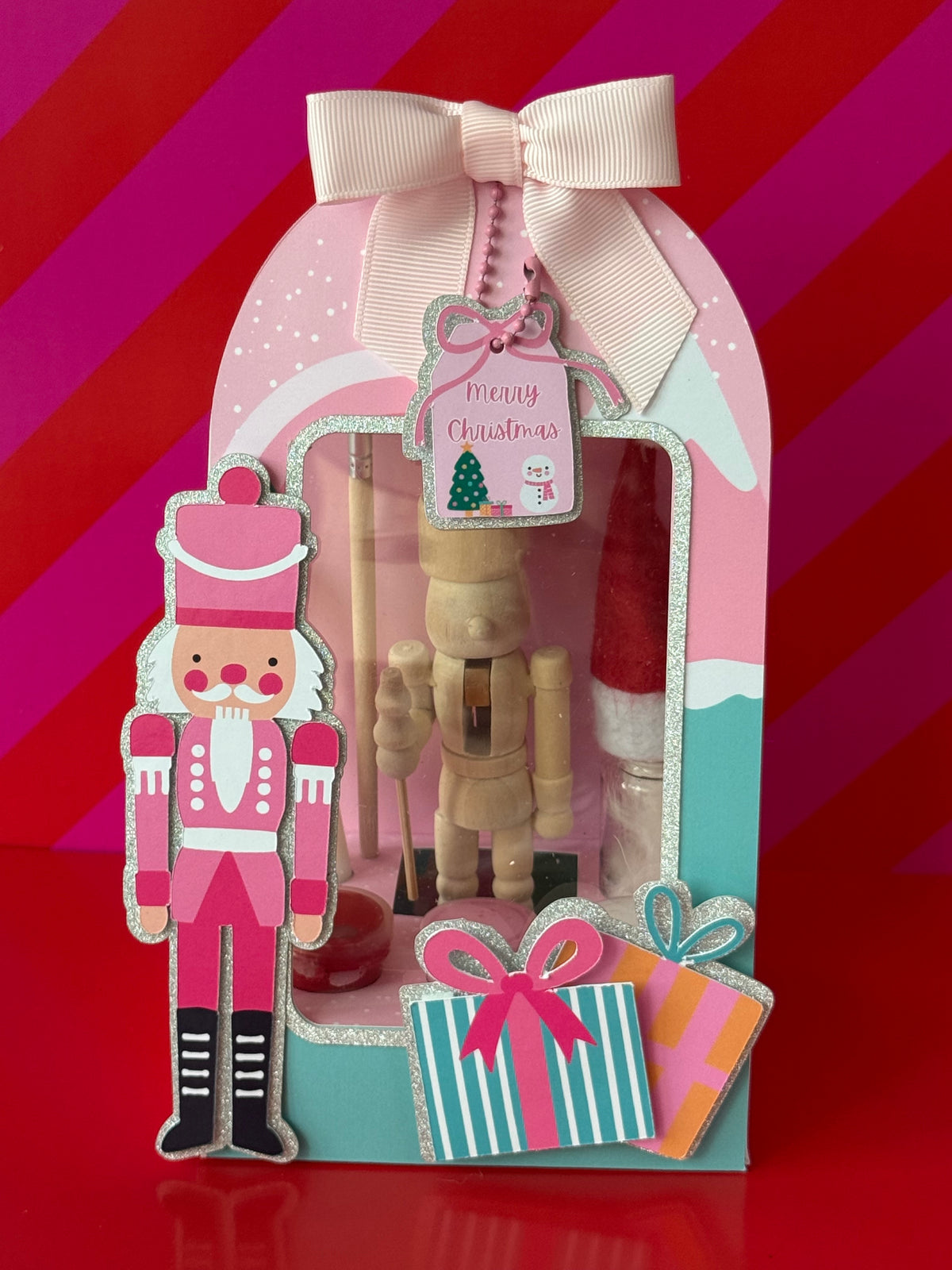 A festive Christmas nutcracker-themed gift set in a decorative arch-shaped box with a large cream ribbon bow. The set includes a pink and silver nutcracker figure, a wooden nutcracker toy, a small red Santa hat, and two wrapped gift boxes with pink bows.