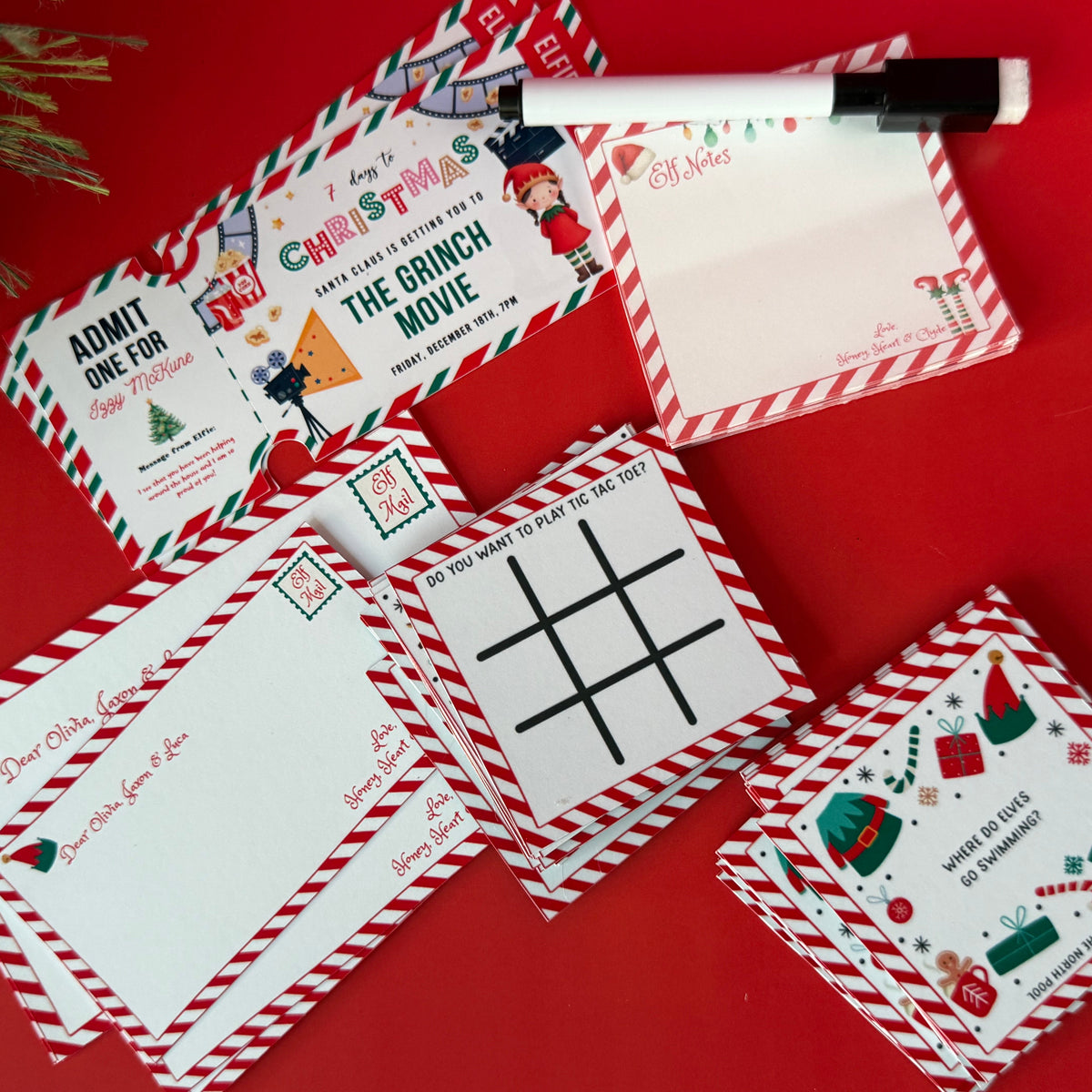 A set of Christmas-themed notepads and envelopes arranged on a bright red background, with a black and white marker resting on top. The stationery features festive red-and-white striped borders and holiday illustrations including Santa Claus, elves, candy canes, and ornaments.
