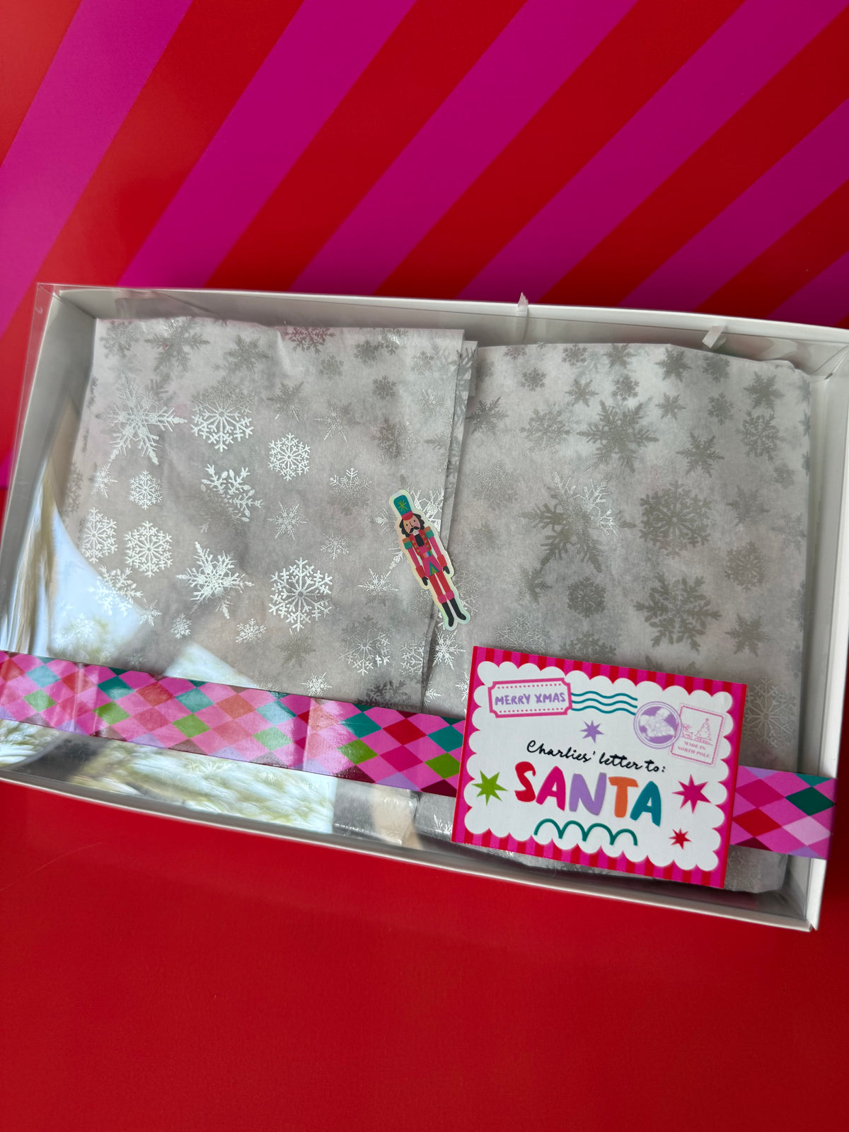 A boxed holiday gift set featuring a silver-gray patterned scarf with snowflakes, a small nutcracker figurine, and a pink and white 'Charities letter to SANTA' card, presented in a clear gift box on a red background.