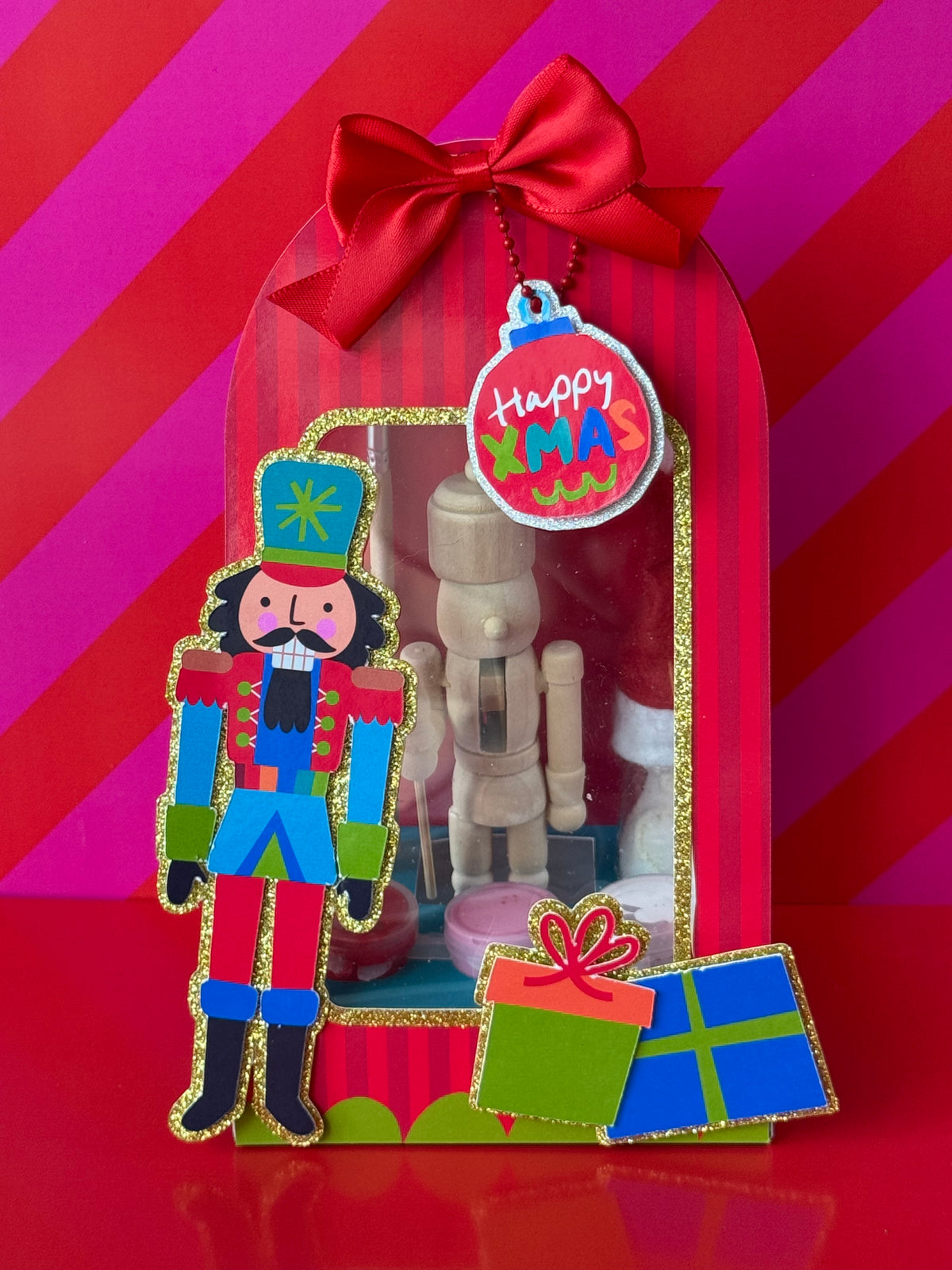A festive Christmas nutcracker-themed gift box set on a red and pink striped background. The set includes a large red bow, a round 'Happy Xmas' ornament tag, a nutcracker figure, a wooden nutcracker, and small gift boxes.