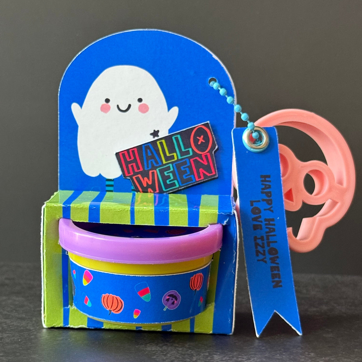 A handmade Halloween-themed paper card holder or gift box with a blue arch back, a white ghost illustration, a striped green and blue base, a purple cup insert, a 'HALLOWEEN' sign, and a blue ribbon tag that reads 'HAPPY HALLOWEEN LOVE 222'. A pink plastic skull keychain is attached on the right, set against a dark gray background.