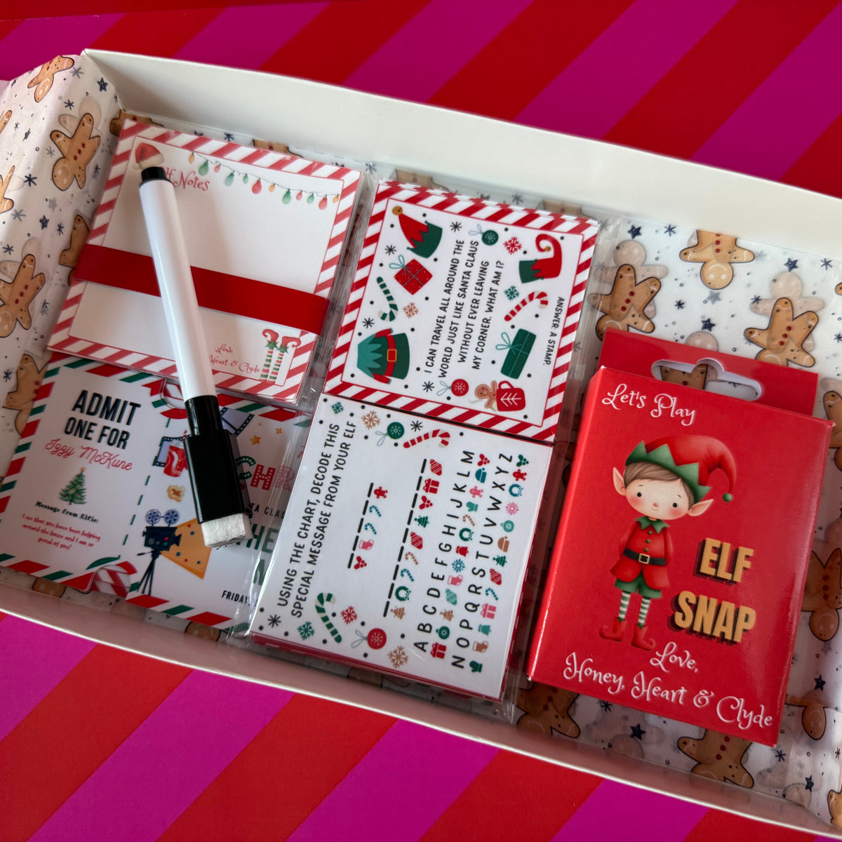 A gift box filled with holiday-themed stationery and a red card. The contents include a white and red striped notepad, a white and red striped calendar sticker sheet, a black and white marker, a gingerbread-patterned notepad, and a red card featuring an elf illustration and the text 'Let's Play Love, Honey Heart & Style'.