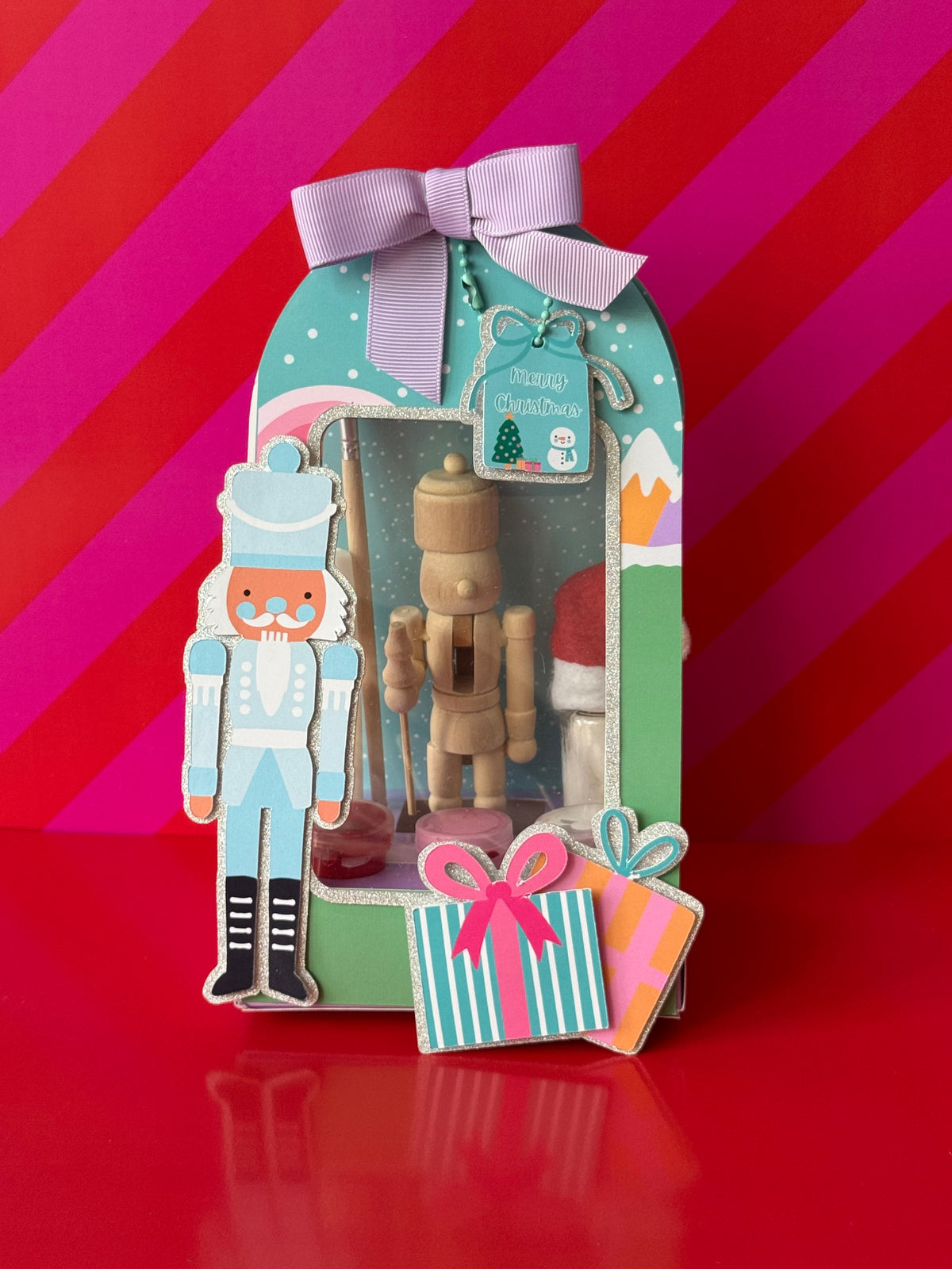 A Christmas-themed gift box set featuring a wooden nutcracker figure inside a decorated teal and green box with a lavender ribbon, alongside a standalone nutcracker and two small wrapped presents on a red surface against a pink striped background.