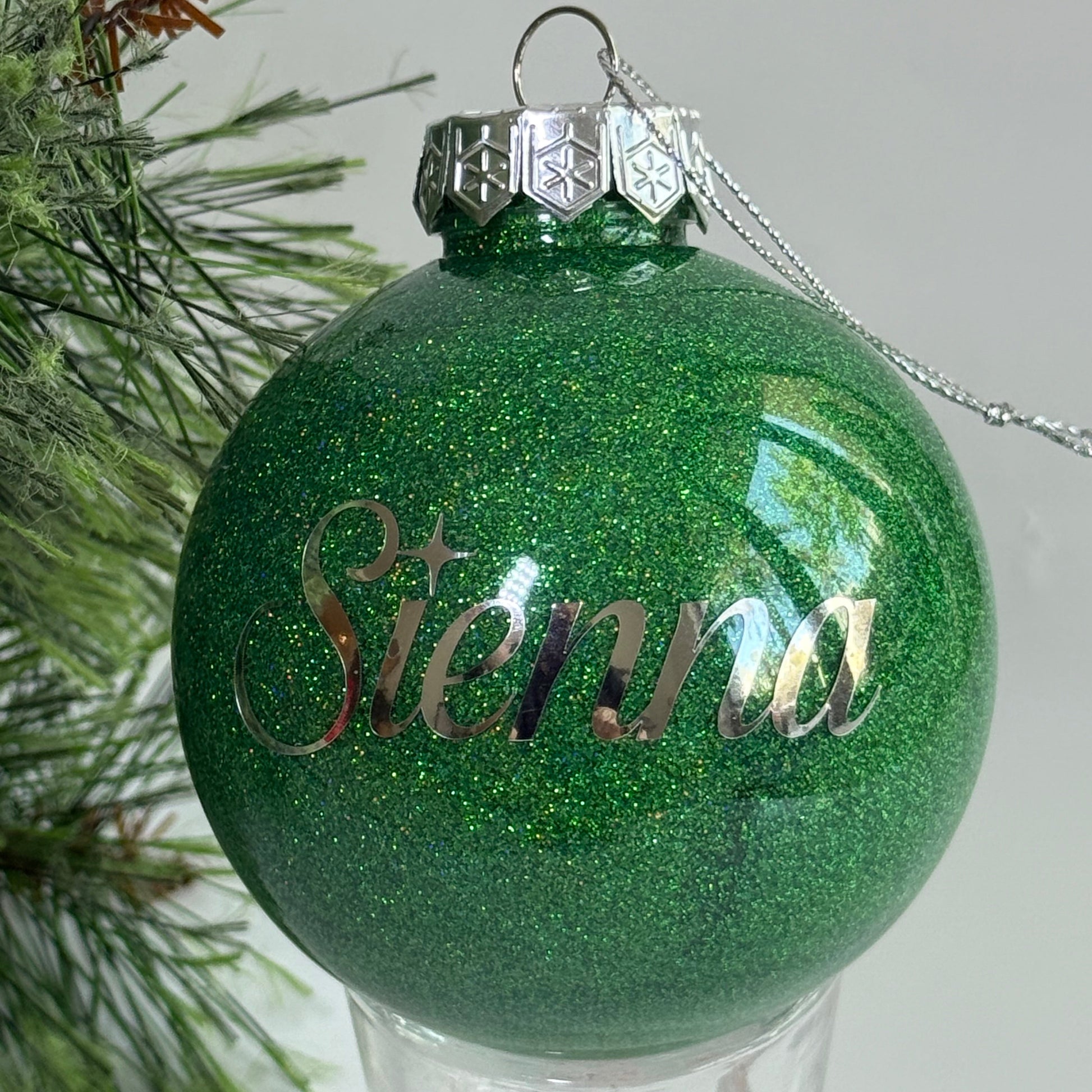 Green glittery Christmas ornament with 'sienna' engraved, hanging on a tree.