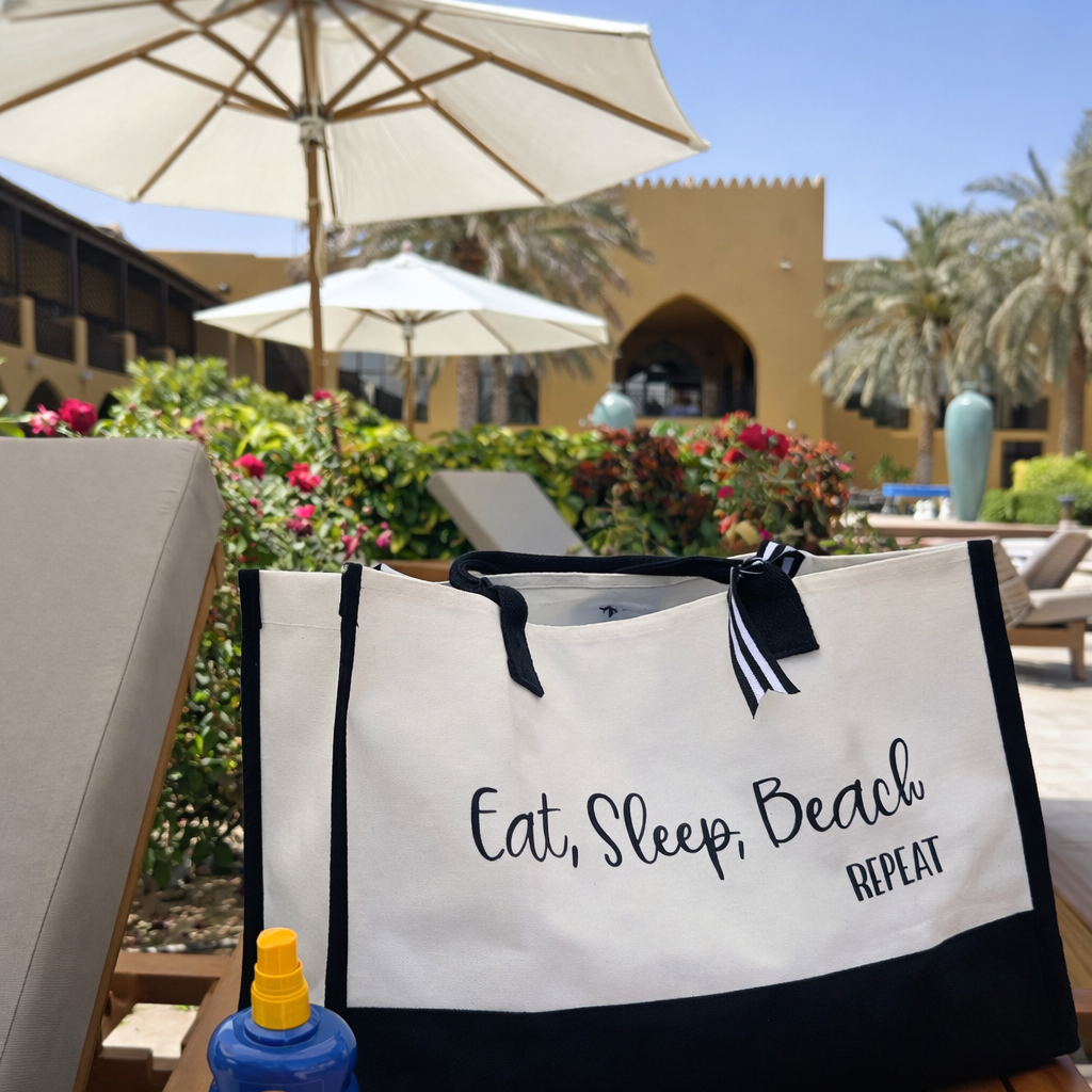A personalized summer tote bag with the text 'EAT SLEEP BEACH' printed in black on the side, displayed at a beach setting with a umbrella and flowers in the background.